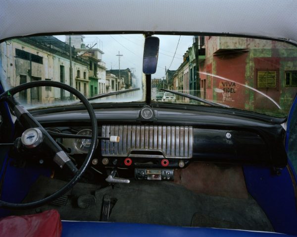 Alex Harris, a car interior looking out along a street, VIVA FIDEL painted on the wall of a building ahead