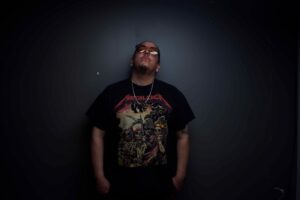 Cloudface, Dine and Hopi artist, photographed against a dark grey wall wearing sunglasses and a black t-shirt.