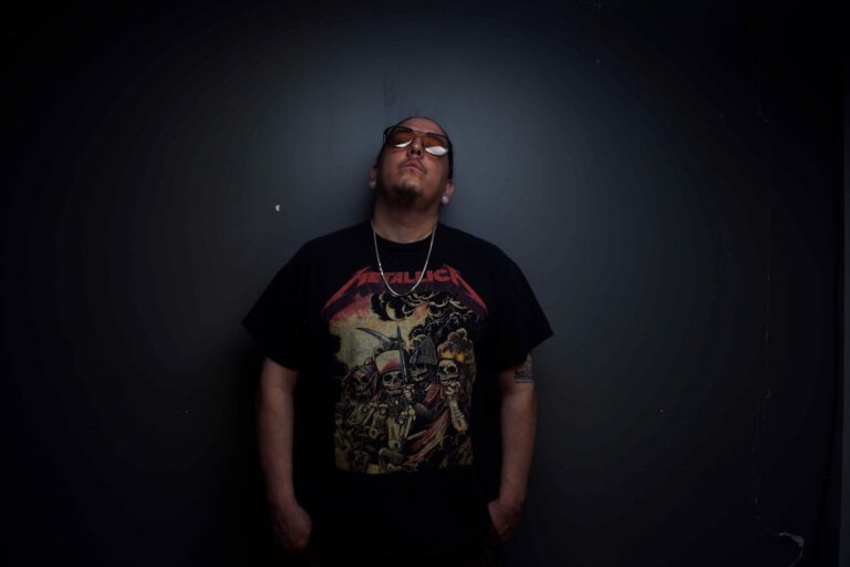 Cloudface, Dine and Hopi artist, photographed against a dark grey wall wearing sunglasses and a black t-shirt.