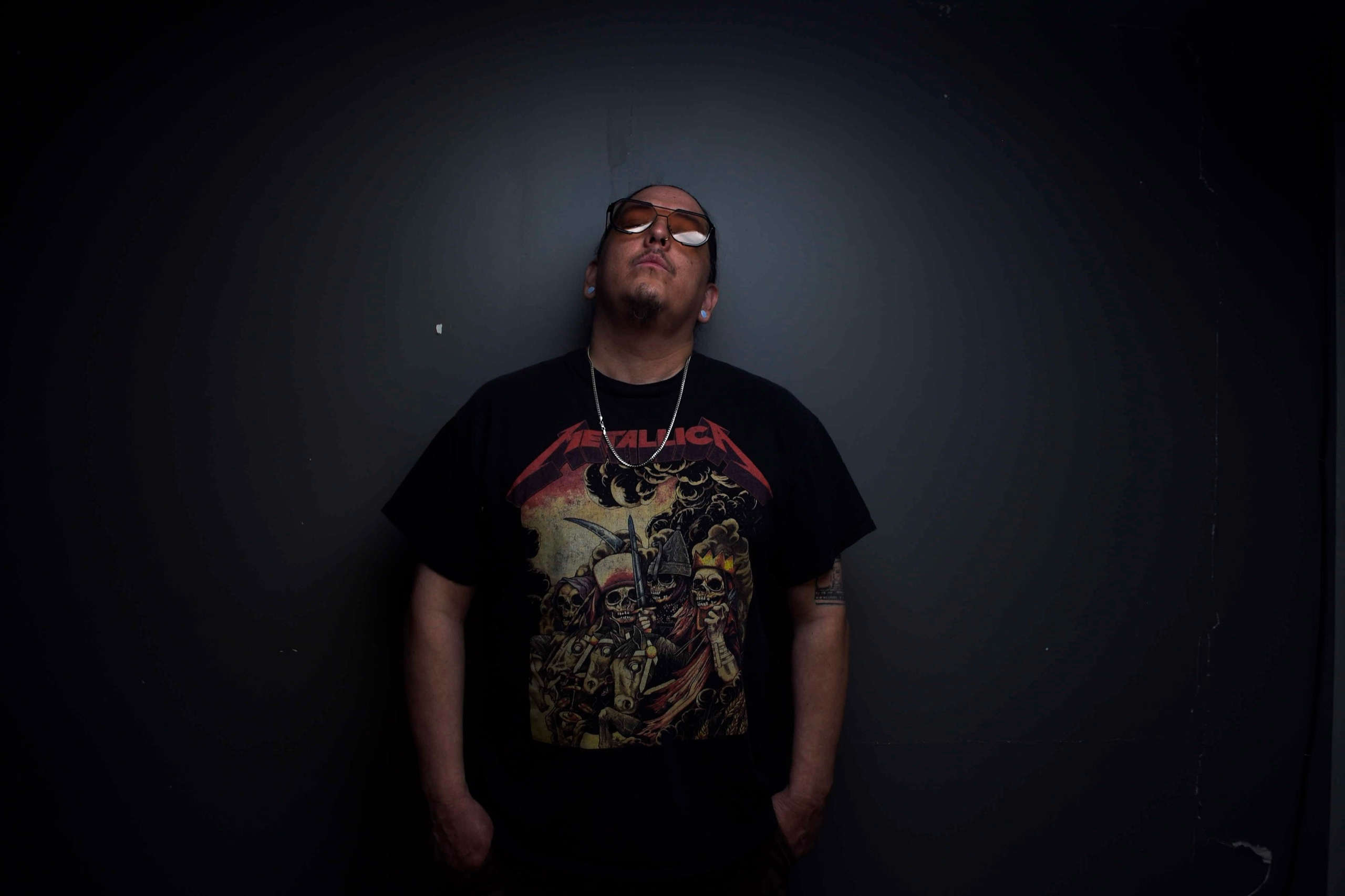 Cloudface, Dine and Hopi artist, photographed against a dark grey wall wearing sunglasses and a black t-shirt.