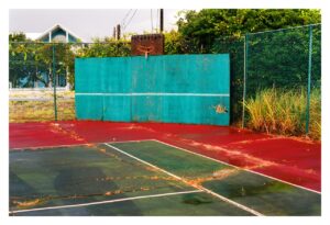 William Greiner, Seacrest Beach, Florida, 2011: a weathered court seen head-on, a teal practice wall with a white stripe and a rusted hoop on a board above it, the cracked red and green surface strewn with orange pine needles, grass through the seams, a blue clapboard house behind