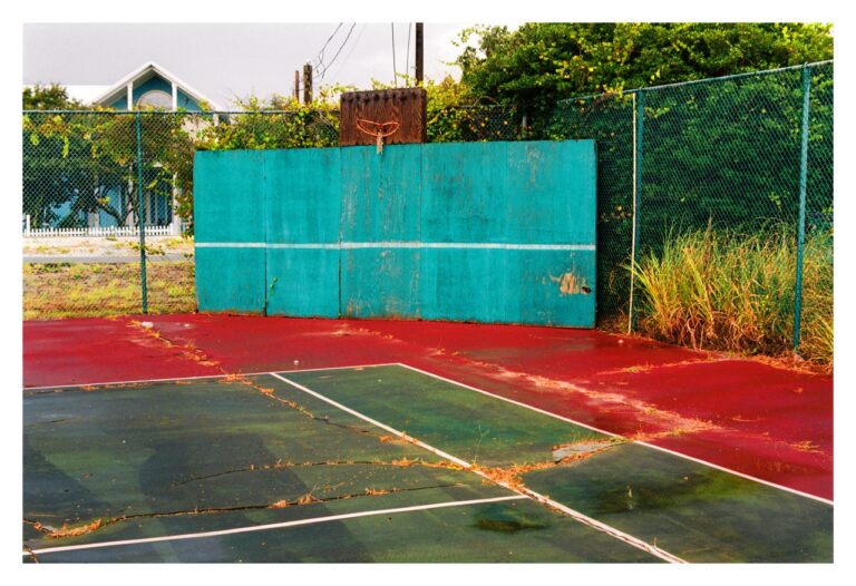 William Greiner, Seacrest Beach, Florida, 2011: a weathered court seen head-on, a teal practice wall with a white stripe and a rusted hoop on a board above it, the cracked red and green surface strewn with orange pine needles, grass through the seams, a blue clapboard house behind
