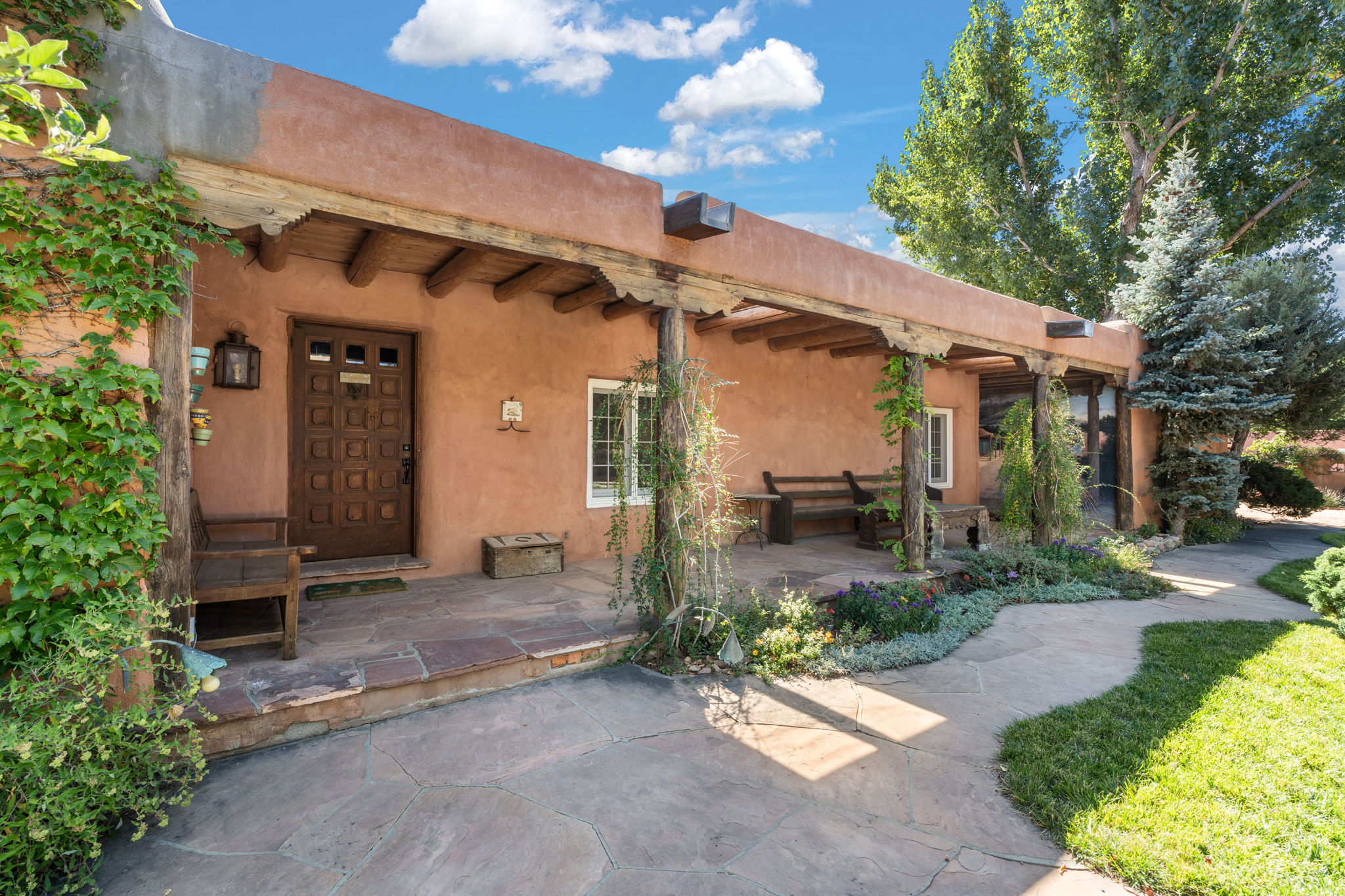 A historic Santa Fe adobe entry on a flagstone courtyard, with viga-supported portal, mature cottonwood, and blue spruce in afternoon light.
