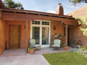 The adobe exterior and portal of a Santa Fe home, with round viga posts, a carved wood door, French doors, stacked firewood, and terracotta pots opening onto a brick courtyard and lawn.