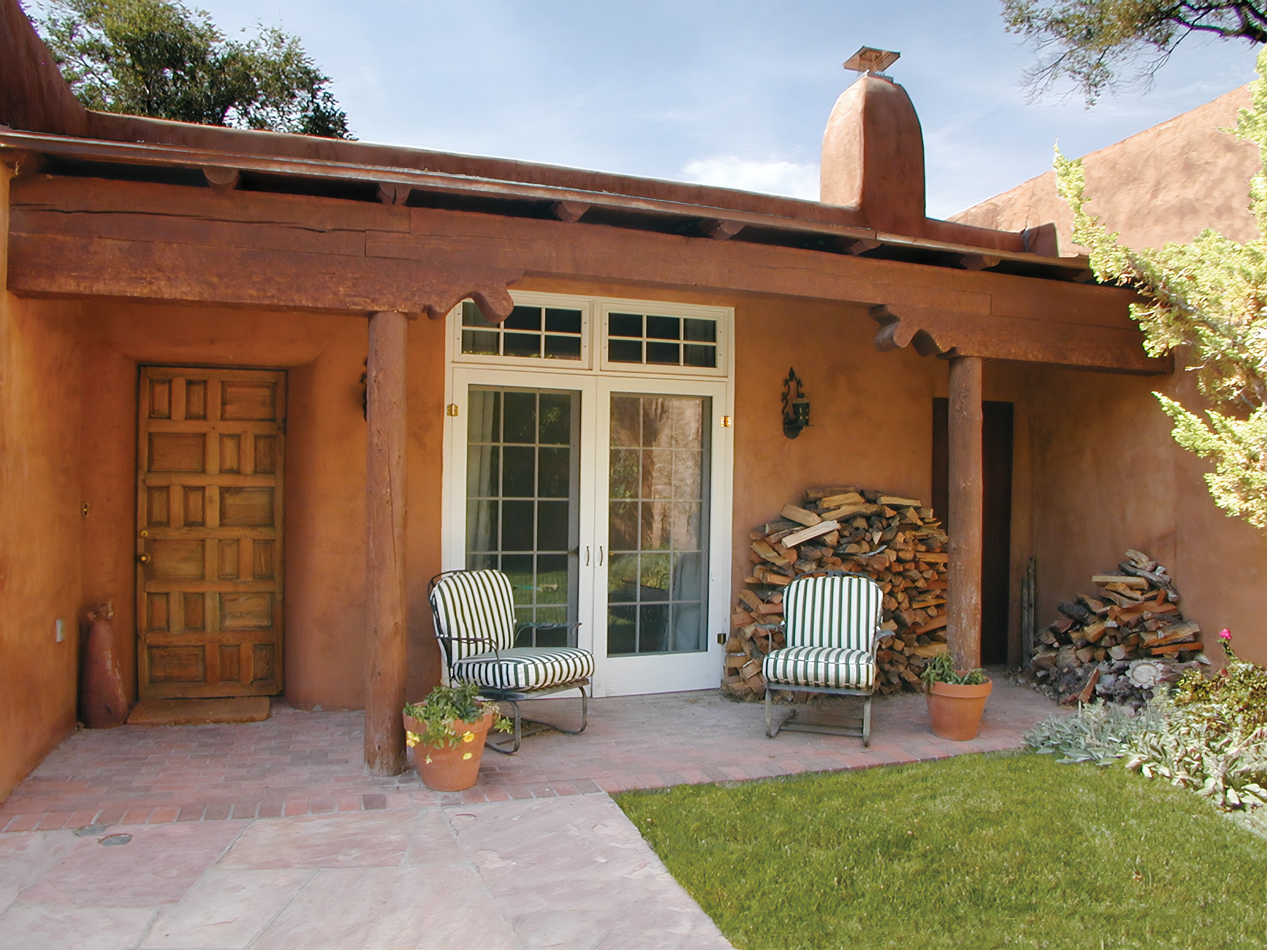 The adobe exterior and portal of a Santa Fe home, with round viga posts, a carved wood door, French doors, stacked firewood, and terracotta pots opening onto a brick courtyard and lawn.