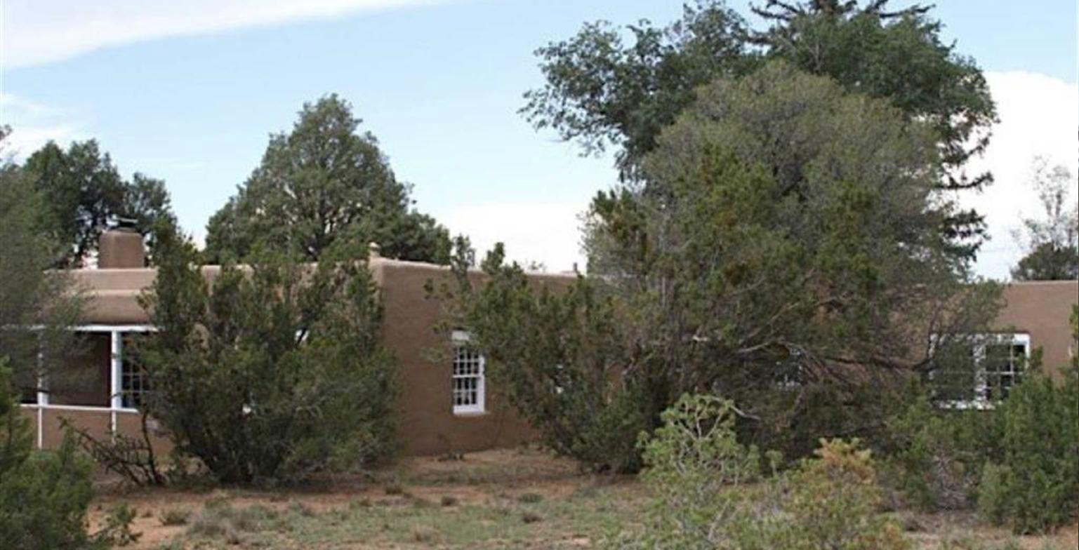 Single story adobe house at 1127 Old Santa Fe Trail with white trimmed windows, a portal, and a chimney behind mature junipers and pinons