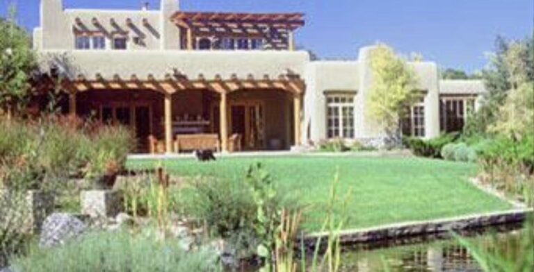 Two story adobe estate at 1422 Bishops Lodge in Tesuque with a long portal, a green lawn, and a pond in the foreground