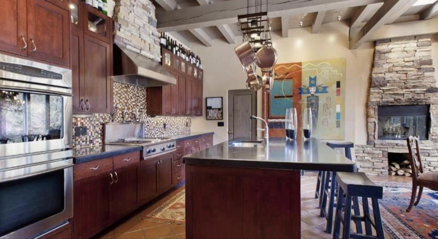 Kitchen at 16 Piedras Negras with cherry cabinets, a stone range hood, a long island with bar stools, and a stacked stone fireplace beyond