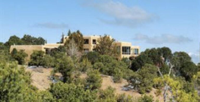 Pueblo style estate at 197 Circle Drive on a wooded hilltop, seen from below through piñon and juniper under a blue sky