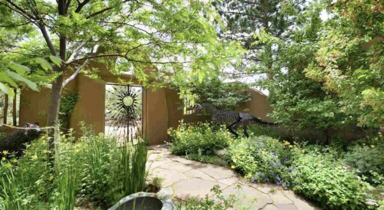 Flagstone path through a flowering garden to an iron gate in an adobe wall with a metal dinosaur sculpture at 29 and 31 Loma Serena