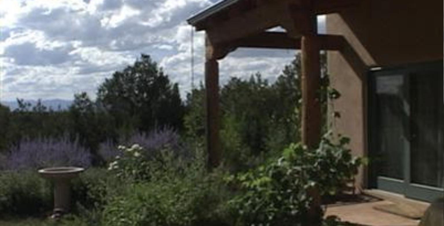 Portal with heavy timber posts at 29 Griego in Tesuque, looking past a birdbath and Russian sage toward piñon hills and the mountains