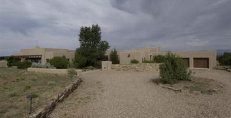Pueblo style house at 3 Golden Eagle in Las Campanas with a stone entry wall, a two car garage, and junipers under a stormy sky