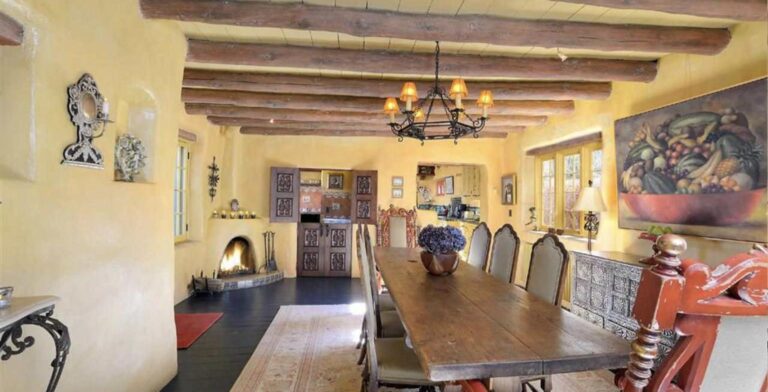 Dining room at 607 Miller with a long wooden table, viga ceiling, iron chandelier, kiva fireplace, and yellow plastered adobe walls