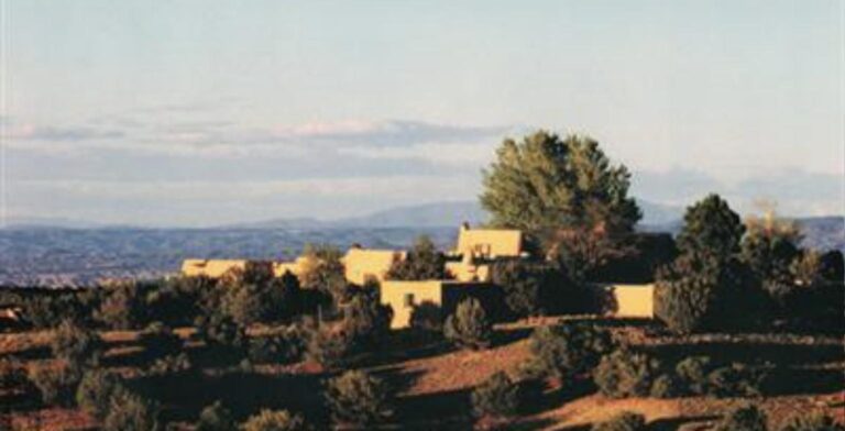 Hilltop adobe compound at 7 Vuelta Susana in La Tierra at sunset, with piñon and juniper below and distant mountains beyond