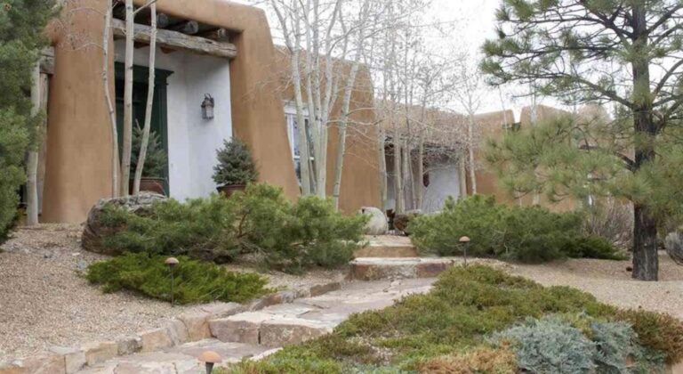 Adobe entry at 708 Camino Militar with a carved lintel, aspens, pines, and a flagstone stair through a landscaped garden