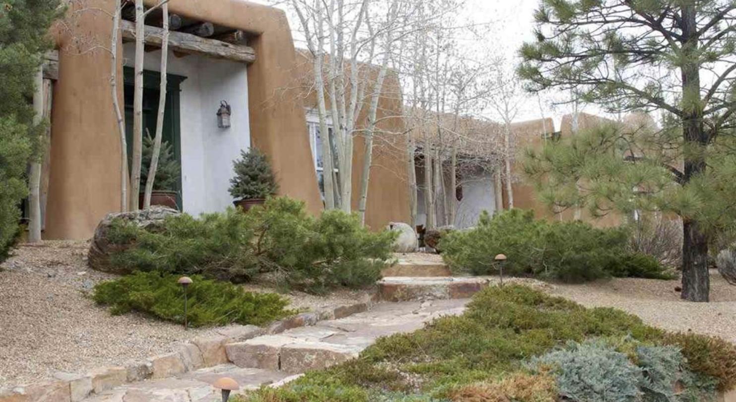 Adobe entry at 708 Camino Militar with a carved lintel, aspens, pines, and a flagstone stair through a landscaped garden