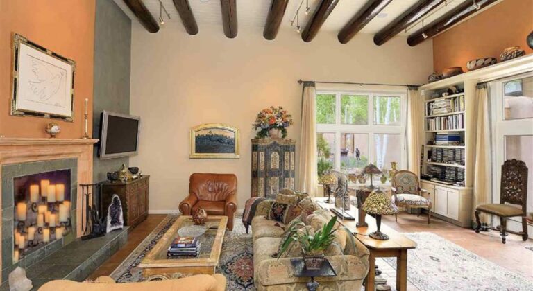Living room at 737 Camino Mirada with a high viga ceiling, a stone fireplace, built in bookshelves, and tall windows onto a garden of aspens
