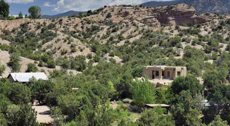 Rancho Rosado at 76 Lavadero in Tesuque, an adobe house with a rooftop deck and a metal roofed studio among piñon and cottonwoods below a sandstone canyon wall
