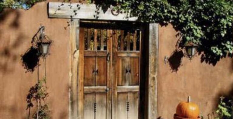 Carved wooden gate in an adobe wall at 793 Camino del Poniente, with iron lanterns, climbing vines, and a pumpkin on the step