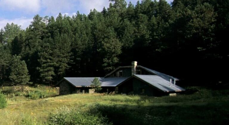 Straw bale house with a pitched steel roof and stone chimney in a mountain meadow below a pine forest at 976 Vallecitos, Jemez Springs