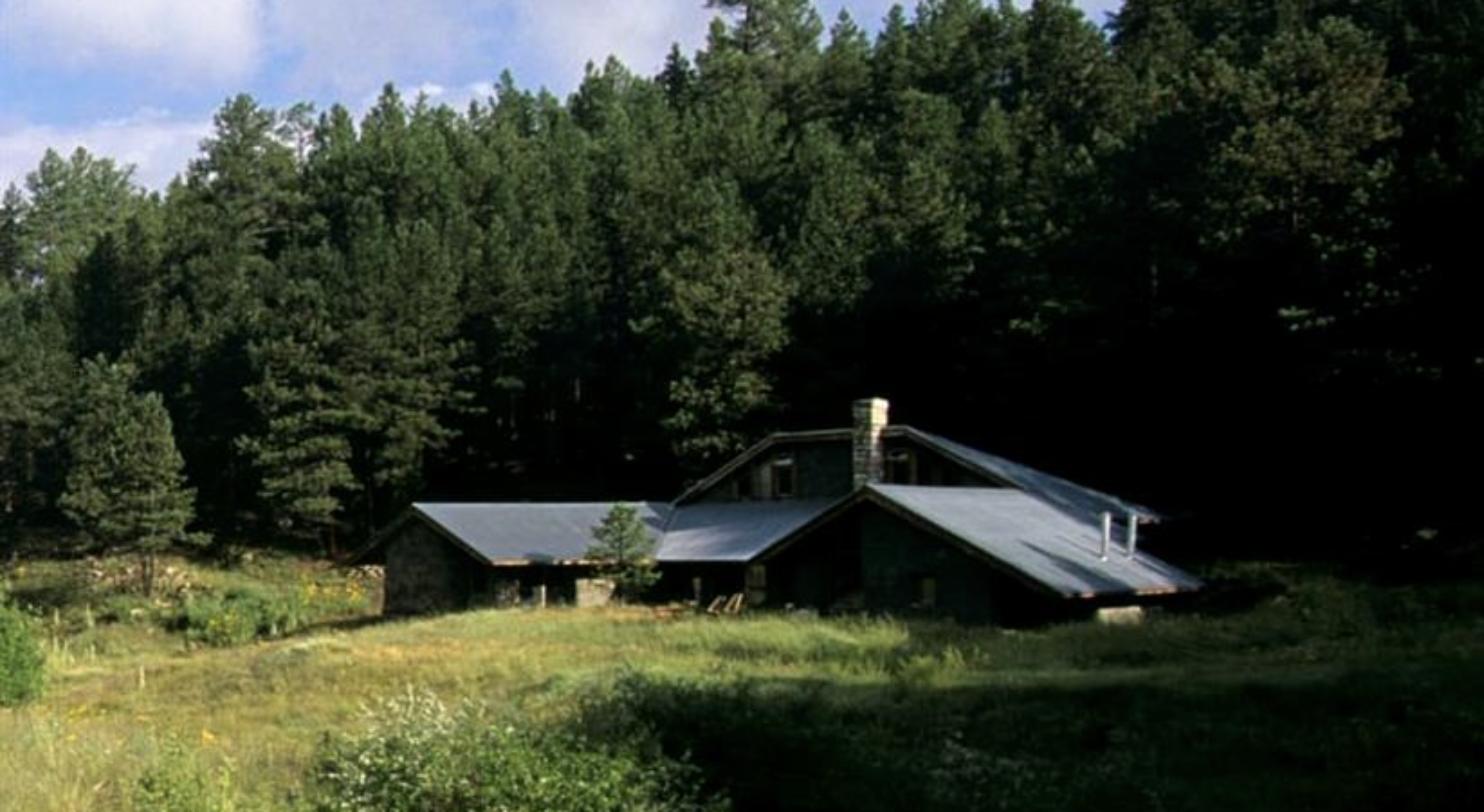 Straw bale house with a pitched steel roof and stone chimney in a mountain meadow below a pine forest at 976 Vallecitos, Jemez Springs