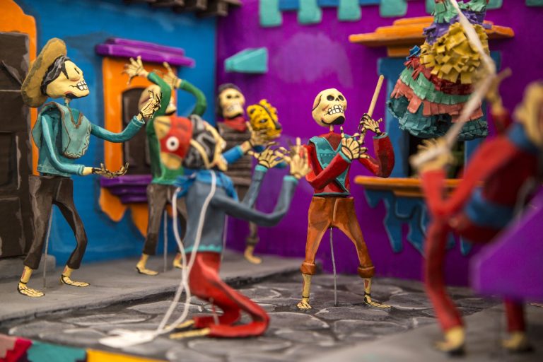 A Day of the Dead tableau of painted skeleton figures in a village street, one swinging a stick at a paper pinata while others dance and play among brightly colored houses.