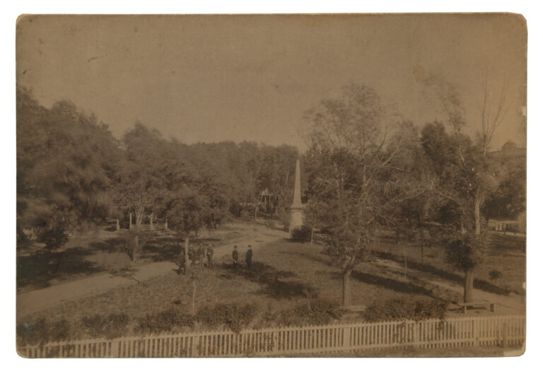 Elevated albumen-print view of the Santa Fe Plaza, 1885: the Soldiers' Monument obelisk at center, five men in hats on the paths, a bandstand at left, and a white picket fence in the foreground.