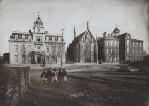 Aged sepia photograph, circa 1880s: the Gothic Revival Loretto Chapel at center with a large traceried rose window, a statue in a gable niche, and a spire behind; the Second Empire Loretto Academy at left with a mansard-roofed tower topped by a cross and the word ACADEMY over its entrance; a tall brick building with a cross-topped cupola at right; and five children in capes standing in a bare dirt lot among leafless trees.
