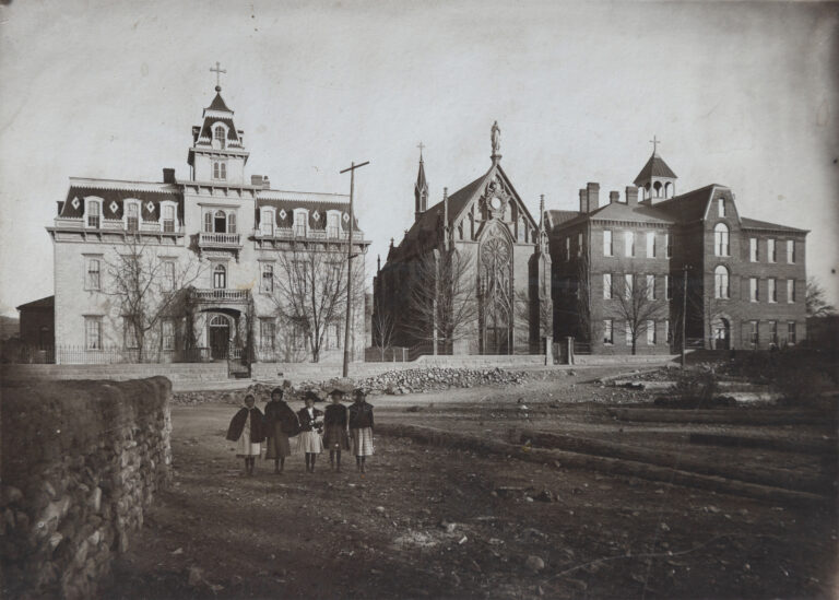 Aged sepia photograph, circa 1880s: the Gothic Revival Loretto Chapel at center with a large traceried rose window, a statue in a gable niche, and a spire behind; the Second Empire Loretto Academy at left with a mansard-roofed tower topped by a cross and the word ACADEMY over its entrance; a tall brick building with a cross-topped cupola at right; and five children in capes standing in a bare dirt lot among leafless trees.