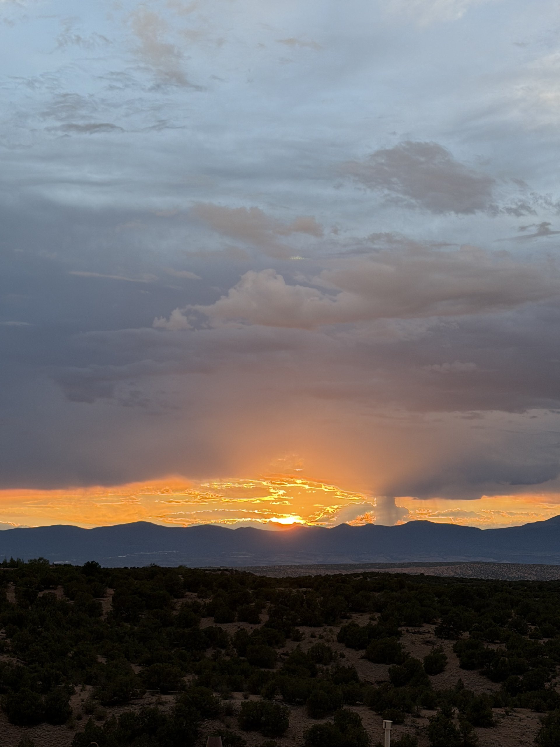 A dramatic sunset over the mountains west of the Santa Fe Opera, the sun dropping through orange clouds above the high-desert pinon, July 24, 2026.
