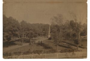 Elevated albumen-print view of the Santa Fe Plaza, 1885: the Soldiers' Monument obelisk at center, five men in hats on the paths, a bandstand at left, and a white picket fence in the foreground.