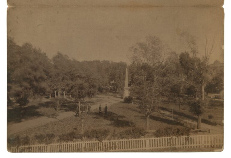 Elevated albumen-print view of the Santa Fe Plaza, 1885: the Soldiers' Monument obelisk at center, five men in hats on the paths, a bandstand at left, and a white picket fence in the foreground.