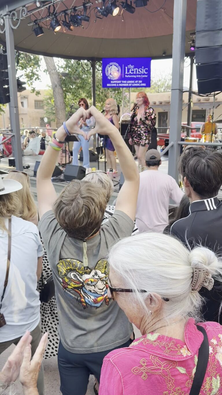 A crowd at the Santa Fe Plaza bandstand at a free Santa Fe Summer Scene concert by the band Abbaquerque, a Lensic Performing Arts Center banner on the stage.
