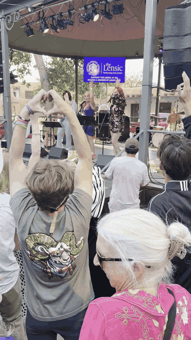 A crowd at the Santa Fe Plaza bandstand at a free Santa Fe Summer Scene concert by the band Abbaquerque, a Lensic Performing Arts Center banner on the stage.