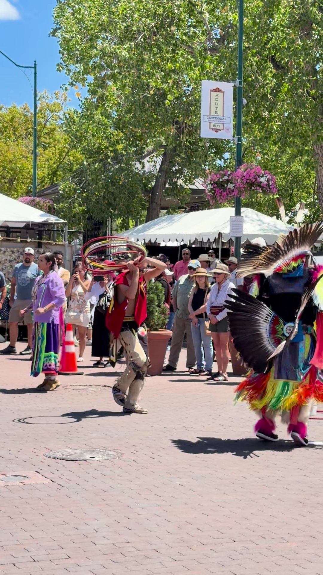 A hoop dancer in regalia raises a bundle of red, yellow, black, and white hoops overhead mid-step on the brick paving of the Santa Fe Plaza during Indian Market, with a crowd in summer hats watching from beneath the cottonwoods and white vendor tents behind.