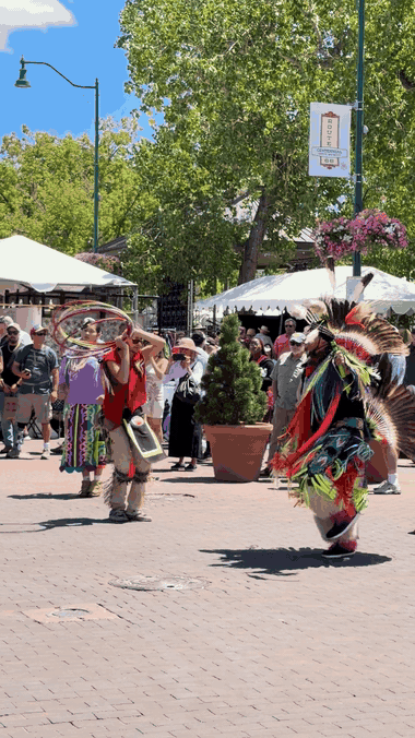 A hoop dancer in regalia turns through a sequence with a bundle of red, yellow, black, and white hoops on the brick paving of the Santa Fe Plaza during Indian Market, a second dancer in a feathered bustle and fringed regalia nearby, as onlookers watch from beneath the cottonwoods in front of the white vendor tents.