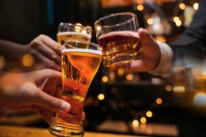 Three hands raising glasses together in a bar, two beers and a tumbler of spirits, with warm lights out of focus behind them.