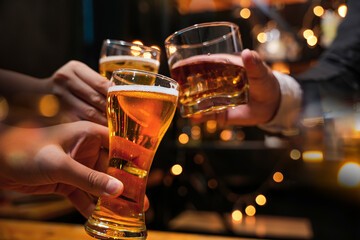 Three hands raising glasses together in a bar, two beers and a tumbler of spirits, with warm lights out of focus behind them.