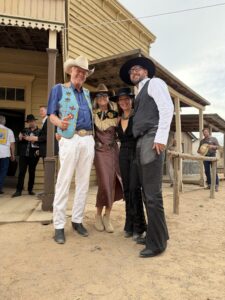 Four guests in Western dress on a covered wooden porch at Bonanza Creek Ranch during the thirty-first Buckaroo Ball, with an adobe wall and other guests behind them.