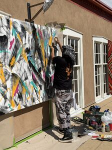 An artist at work on the Carrier Wave canvas on the rooftop at Webster Collection during Indian Market weekend, kneeling over a large painting of black brushwork with yellow, teal, and magenta marks, spray cans and rollers set out beside it, gallery windows and a Navajo textile on the wall behind.
