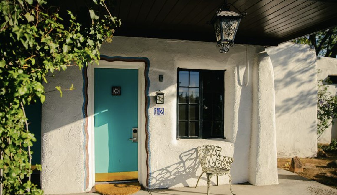 A guest room door at El Rey Court, painted turquoise inside a scalloped border on a white adobe wall, with the room number beside it, an iron lantern hanging from the portal, and vines across the frame in late afternoon light.