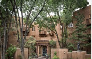The courtyard entrance of Hotel Santa Fe, a stepped adobe building with wooden balconies and window surrounds, seen through mature cottonwoods, with a bronze figure standing among planted beds to the right of the doors.