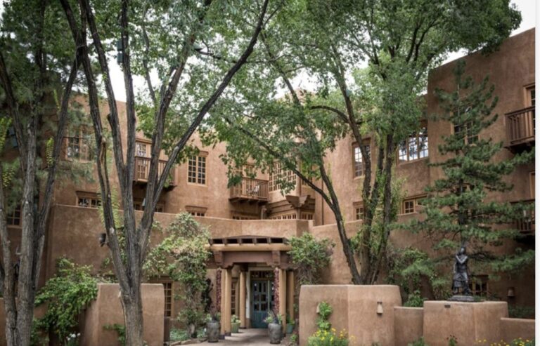 The courtyard entrance of Hotel Santa Fe, a stepped adobe building with wooden balconies and window surrounds, seen through mature cottonwoods, with a bronze figure standing among planted beds to the right of the doors.