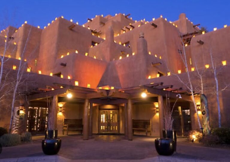 The Inn and Spa at Loretto at dusk, its terraced adobe massing outlined with rows of lit farolitos, the timber portal over the entrance glowing beneath, and bare aspens on either side against a deep blue sky.
