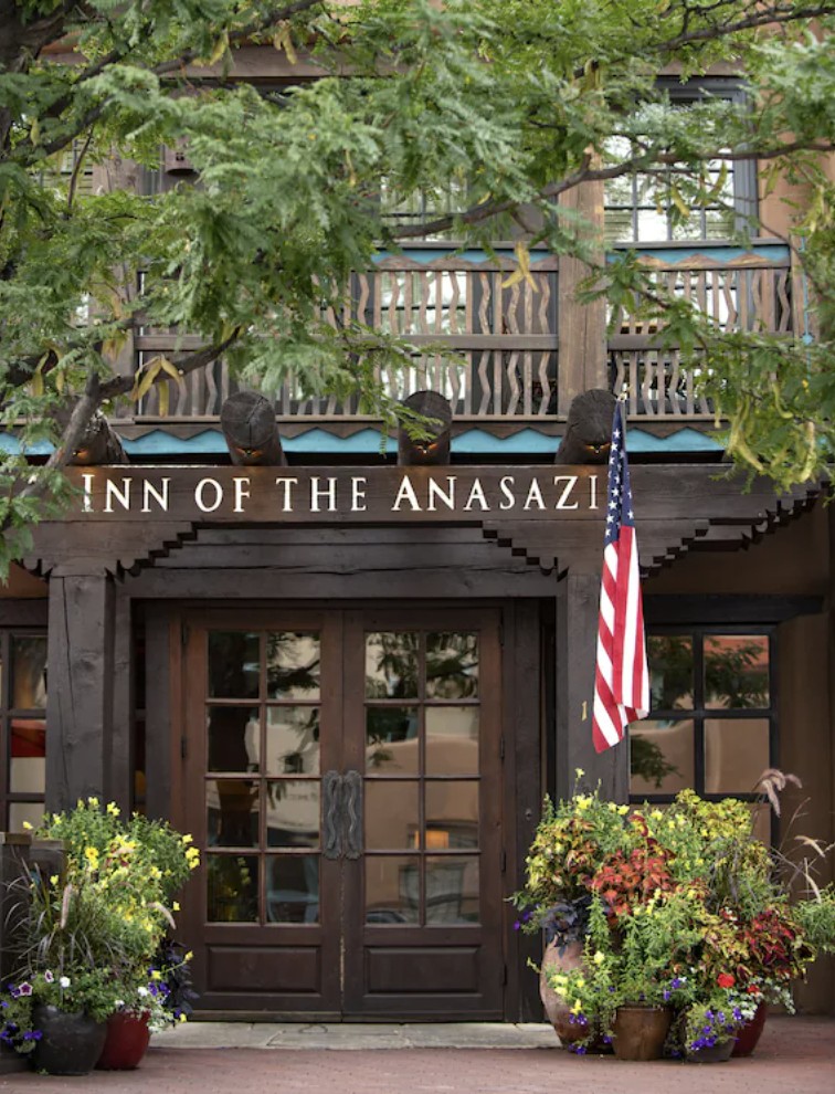 The entrance to the Inn of the Anasazi on Washington Avenue, a heavy timber portal over carved wooden doors with the hotel name in raised letters above them, carved figures along the beam, and planters of flowers on either side.