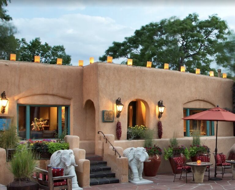 The adobe front of the Inn of the Five Graces at dusk, farolitos burning along the parapet, carved stone elephants flanking the steps, and small tables set out on the flagstone courtyard.