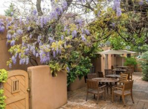 A courtyard at the Inn on the Alameda with wisteria in flower spilling over an adobe wall above a carved wooden gate, and teak tables and chairs set out on brick paving under a canvas umbrella.