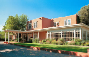 A John Gaw Meem house on Old Pecos Trail, its two storey adobe mass in salmon plaster set behind a white painted portal, with a long glazed conservatory running along the front and planted borders against the lawn.