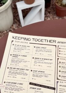 The printed beer list at Keeping Together, lying on a dark round patio table in the beverage garden, with potted plants and a low white stool on the gravel behind it.