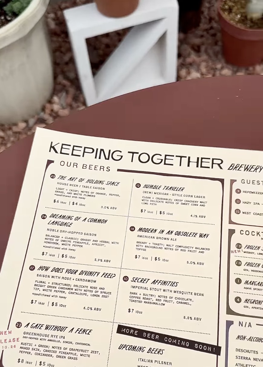 The printed beer list at Keeping Together, lying on a dark round patio table in the beverage garden, with potted plants and a low white stool on the gravel behind it.